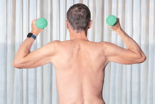 Shirtless senior man seen from rear and exercising with dumbbells in bedroom against curtains