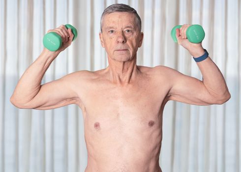 Shirtless senior man seen from front and exercising with dumbbells in bedroom against curtains