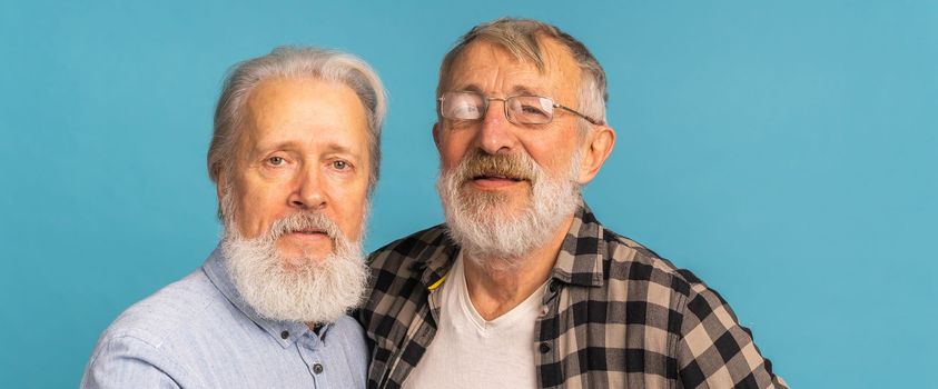 Portrait two elderly man friends standing over blue background - friendship, aged and senior