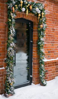 Stylish christmas fir branches with red baubles and sparkling garland on front of door at holiday market or restaurant in city street. Winter christmas street decor