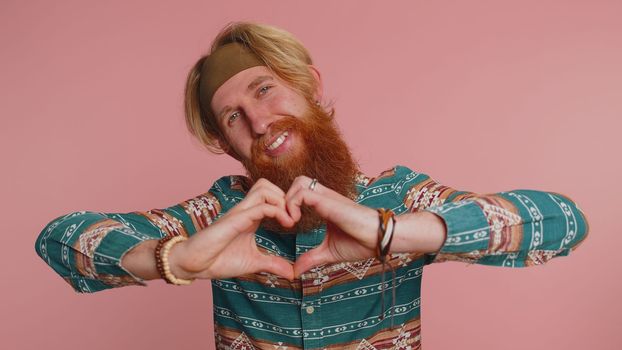 Man in love. Smiling hippie man in pattern shirt makes heart gesture demonstrates love sign expresses good feelings and sympathy. Young redhead adult guy boy isolated alone on pink studio background