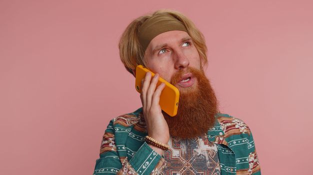 Tired sleepy freelancer hippie redhead bearded man talking on mobile phone with friend making online conversation. Disinterested hipster guy having annoyed boring talk on smartphone on pink background