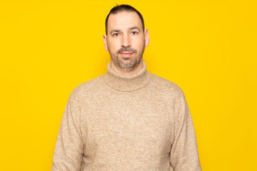 Handsome bearded hispanic man wearing beige turtleneck sweater posing smiling for studio portrait over yellow background