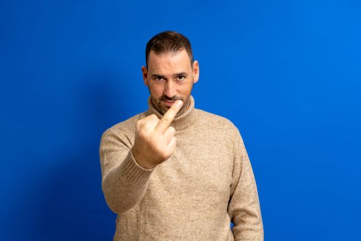 Bearded hispanic man wearing pullover standing over isolated blue background. Showing middle finger, impolite and rude shitty expression