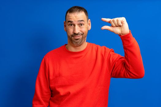 Hispanic man standing over blue background smiling and confident gesturing with hand making little sign with fingers looking and camera. measure concept.