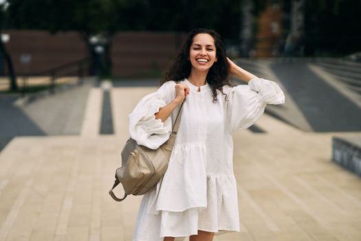 Smiling curly brunette girl in a white dress with a backpack putting her hand behind her head. High quality photo