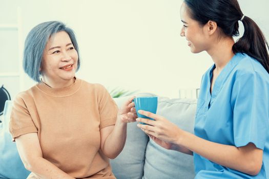 Female care taker serving her contented senior patient with a cup of coffee at home, smiling to each other. Medical care for pensioners, Home health care service.
