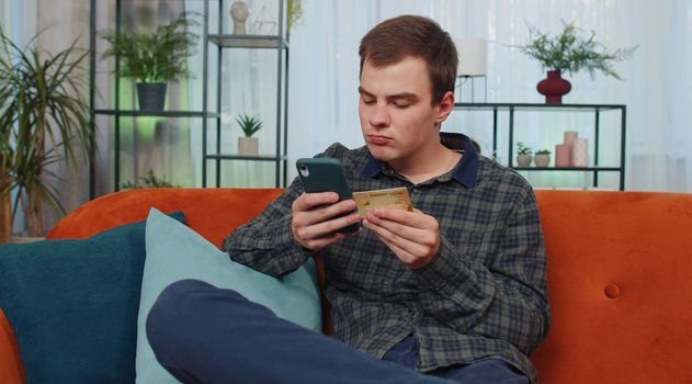 Teenager man using credit bank card and smartphone while transferring money, purchases online shopping app, order food delivery at home apartment indoors. Young guy in living room sitting on couch