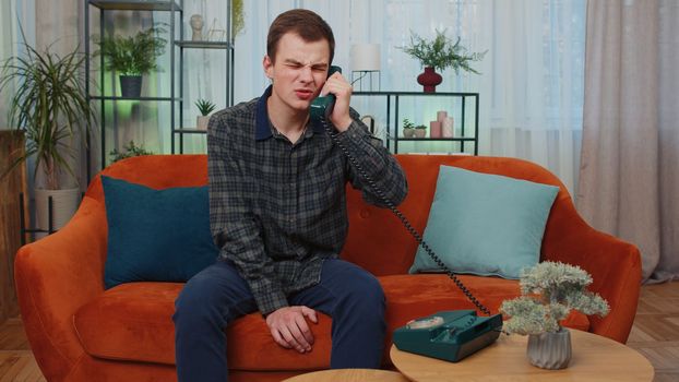 Portrait of teenager man making wired telephone conversation with friends sitting call on couch at home in room. Happy excited young guy enjoying old-fashioned retro phone from 90s talking indoors