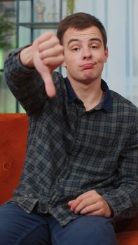 Dislike. Upset teen man showing thumbs down sign gesture, expressing discontent disapproval dissatisfied bad loser work at modern home apartment indoor. Displeased guy in living room on sofa. Vertical