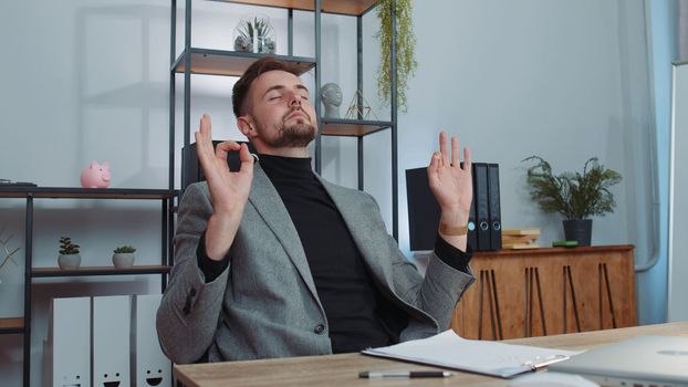 Businessman in suit working on laptop computer, meditating, doing yoga breathing exercise in lotus position at home office desk. Calm serene freelancer man taking break. Busy occupation. Peace of mind