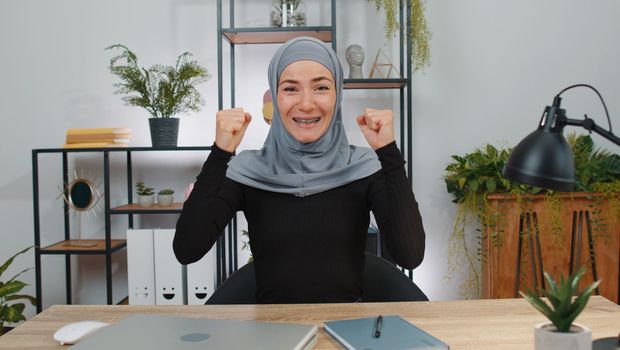 Happy young muslim business woman wearing hijab headscarf shouting, celebrating success victory, winning, goal achievement, good news, lottery jackpot luck, surprise. Girl at home office workspace