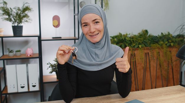 Young muslim business woman wearing hijab real estate agent lifting hand showing the keys of new home house apartment, buying or renting property, mortgage loan. Girl at modern home office workspace