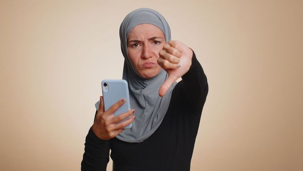Muslim woman in hijab use mobile cell phone typing browsing, loses becoming surprised sudden lottery results, bad fortune, loss, fail. Young arabian girl isolated alone on beige studio wall background