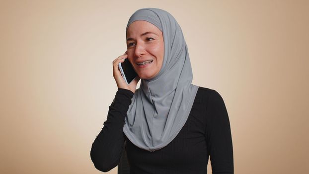 Happy young muslim woman in hijab making call using mobile cell phone, talking pleasant conversation with good friend or family. Arabian female girl speaking on smartphone on studio beige background