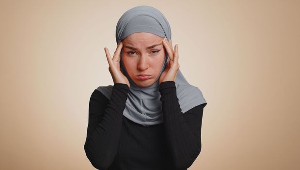 Unhealthy young muslim woman in hijab rubbing temples to cure headache problem, suffering from tension, migaine, stress, grimacing in pain, high blood pressure isolated alone on beige wall background
