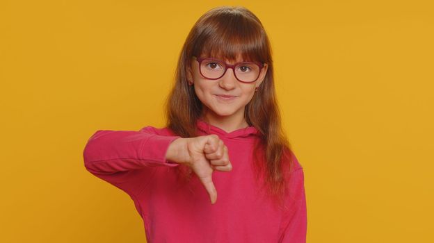 Dislike. Upset young preteen child girl kid showing thumbs down sign gesture, expressing discontent, disapproval, dissatisfied bad work indoors. Displeased toddler children on yellow studio background