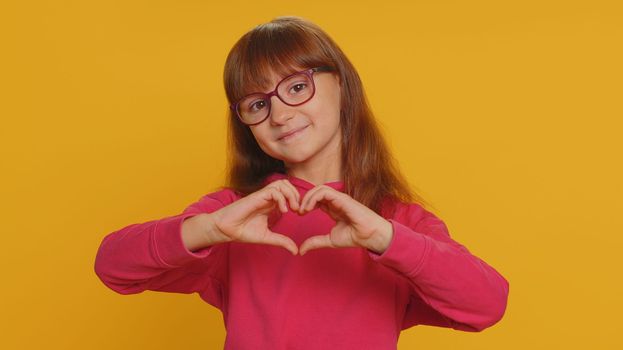 I love you. Smiling young preteen child girl kid makes heart gesture demonstrates love sign expresses good feelings and sympathy. Little toddler children isolated alone on studio yellow background