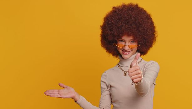Happy funny girl showing thumbs up pointing empty place, advertising area for commercial text, copy space for goods promotion. Young little child kid. Studio shot indoors on yellow wall background
