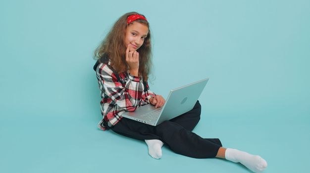 Happy young teenager child girl kid 12 years old works on notebook laptop pc, sends messages, makes online purchases, watching movies. Preteen children isolated sitting on studio blue background