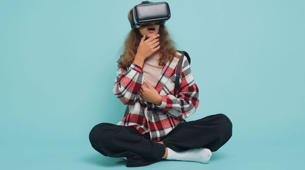 Teenager child girl kid using virtual reality futuristic technology VR app headset helmet to play simulation 3D 360 video game, watching film movie. Preteen children in goggles sits on blue background