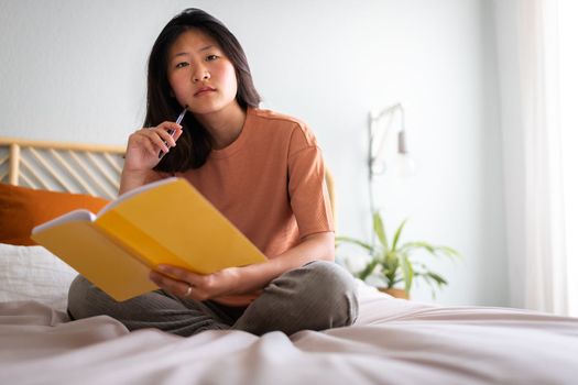 Serious, pensive teen asian female college student sitting on bed writing on diary looking at camera. Copy space. Lifestyle concept.