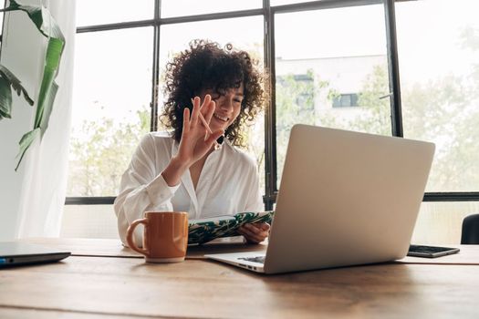 Young mixed race female sitting behind desk waving a coworker in an online work meeting. Working from home concept. Technology concept.