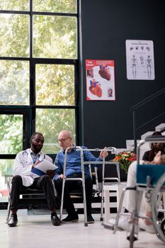 Practitioner doctor holding clipboard with disease diagnosis results explaining medical expertise to senior patient with wallking frame during checkup visit in hospital waiting area. Medicine service