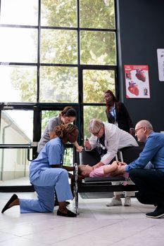 Family being worried after unconscious caucasian kid falling down on chair in hospital waiting area during examination. Medic consulting unconscious little child shining light into eyes.