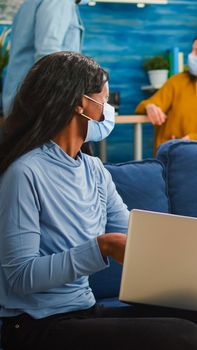 Happy cheerful woman holding beer bottle looking at laptop talking with african friend in living room wearing face mask to prevent coronavirus spread in time of global pandemic. Conceptual footage.