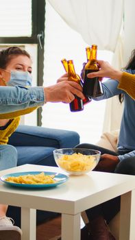 Happy diverse young people having fun in living room, clinking bottles of beer telling stories and jokes during global pandemic. Multiethnic group of friends celebrating with toast in outbreak