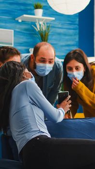 African woman showing funny clip on smartphone hanging out in home living room wearing face mask preventing coronavirus during global pandemic keeping social distancing, enjoying party in outbreak