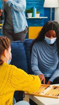 African woman competing with friends playing backgammon wearing face mask keeping social distancing during social pandemic with covid19 drinking beer in living room. Enjoying board games in outbreak