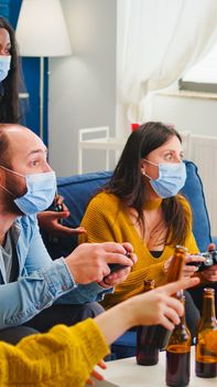 Sad friends because loosing playing video games with wireless controller wearing face mask to prevent getting sick with corona in time of social pandemic. Group of people enjoying time together.