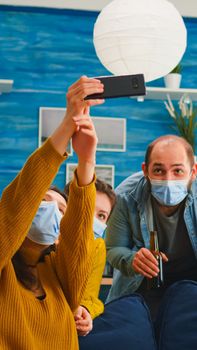 Cheerful happy mixed race friends taking selfie wearing face mask keeping social distancing against spread of virus sitting on couch in living room. Diverse people socializing keeping social distance