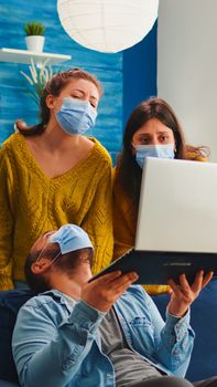Group of mixed race friends having fun keeping social distancing with face mask preventing coronavirus spreading looking at video on laptop. in living room drinking beer and eating chips.
