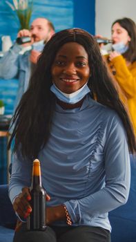 African woman keeping social distancing wearing face mask while meeting with friends to prevent spread of coronavirus holding beer bottle looking at camera sitting on couch, covid 19 outbreak