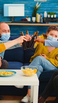 Diverse group of friends celebrating with toast clinking raised bottles of beer keeping social distancing to prevent coronavirus spread. during global pandemic having fun in home living room.