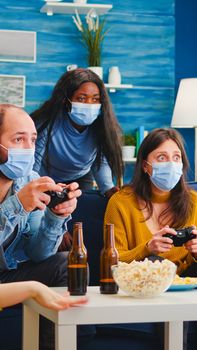 Concentrated multiracial friends playing video games with joystick controller in living room wearing face mask to prevent spreading coronavirus. Diverse people having fun, laughing at new normal party
