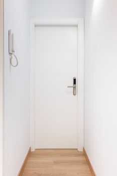 White entrance door to an apartment with metal handle and an electronic lock in center of white bright hallway with parquet flooring. On wall is tube from intercom in entrance
