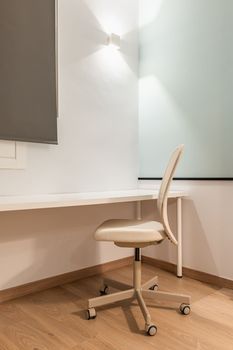 Close-up of work area with white table against wall with beige soft leather office chair for comfortable and productive work online on parquet floor. Room with bright artificial light