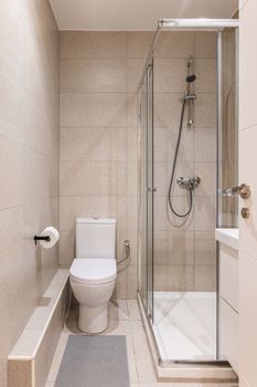 Entrance to a small bathroom with beige marble tiles on the walls and floor, white clean toilet, vanity sink, glass-enclosed shower area. Bathroom is illuminated by light bulbs on the ceiling