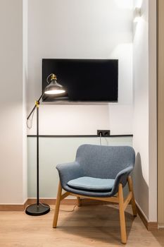 Closeup of modern comfortable armchair with dark blue upholstery bright ceiling lights and metal floor lamp in room with white walls and wooden parquet. Behind TV on wall for watching shows