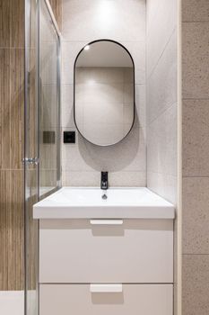 Closeup of a bathroom alcove with bright artificial lighting, with a white solid ceramic sink on a two-drawer vanity. Large modern oval mirror on the wall above the sink