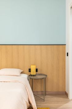 Vertical view of a bed with pillow with large warm duvet and linens in pleasant beige color. Modern design wall with wood paneling. On table is lamp with yellow frame for soft warm light at night