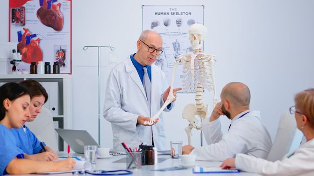 Senior man doctor showing the work of human's hand on skeleton anatomical model. Specialist medic explaining diagnosis to colleagues standing in front of desk in hospital meeting room.