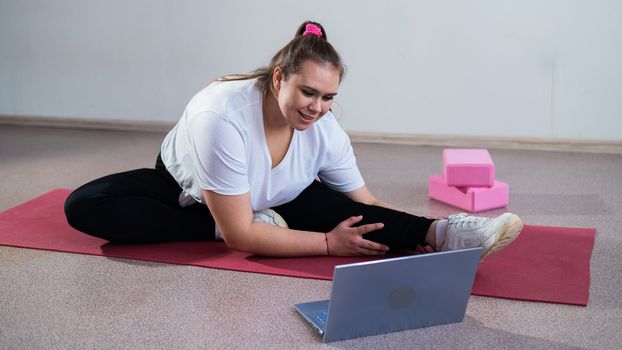 Young caucasian fat woman doing bends on a sports mat and watching a training video on a laptop. A chubby girl doing stretching remotely using video communication.