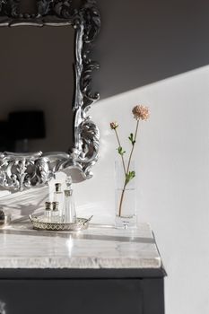 Part of dressing table with vase and flower bright by sunlight on white marble top. Mirror in silver frame with beautiful figured monograms. On table are empty bottles of fragrant perfume