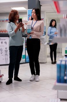 Client showing pills on smartphone to specialist, asking for pharmaceutical products to buy at drugstore. Elderly woman and consultant looking at medical supplements and drugs box.