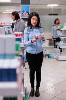 Asian customer reading pills box leaflet before buying healthcare products from drugstore shelves. Pharmacy client looking at medicaments and vitamins packages, cardiology drugs.